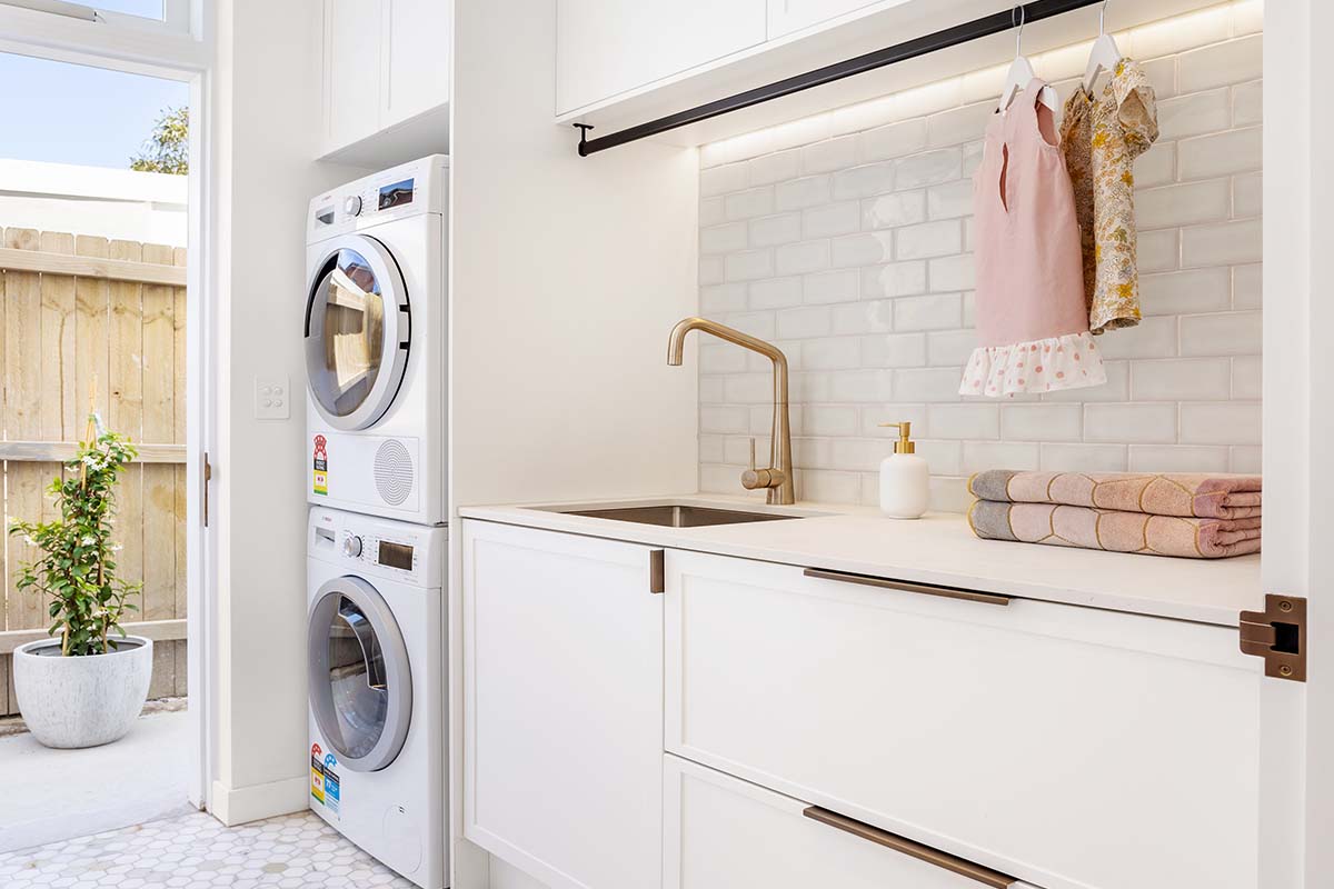 White laundry with stacked washer and dryer, hanging rail, tiled splashback and brass tapware