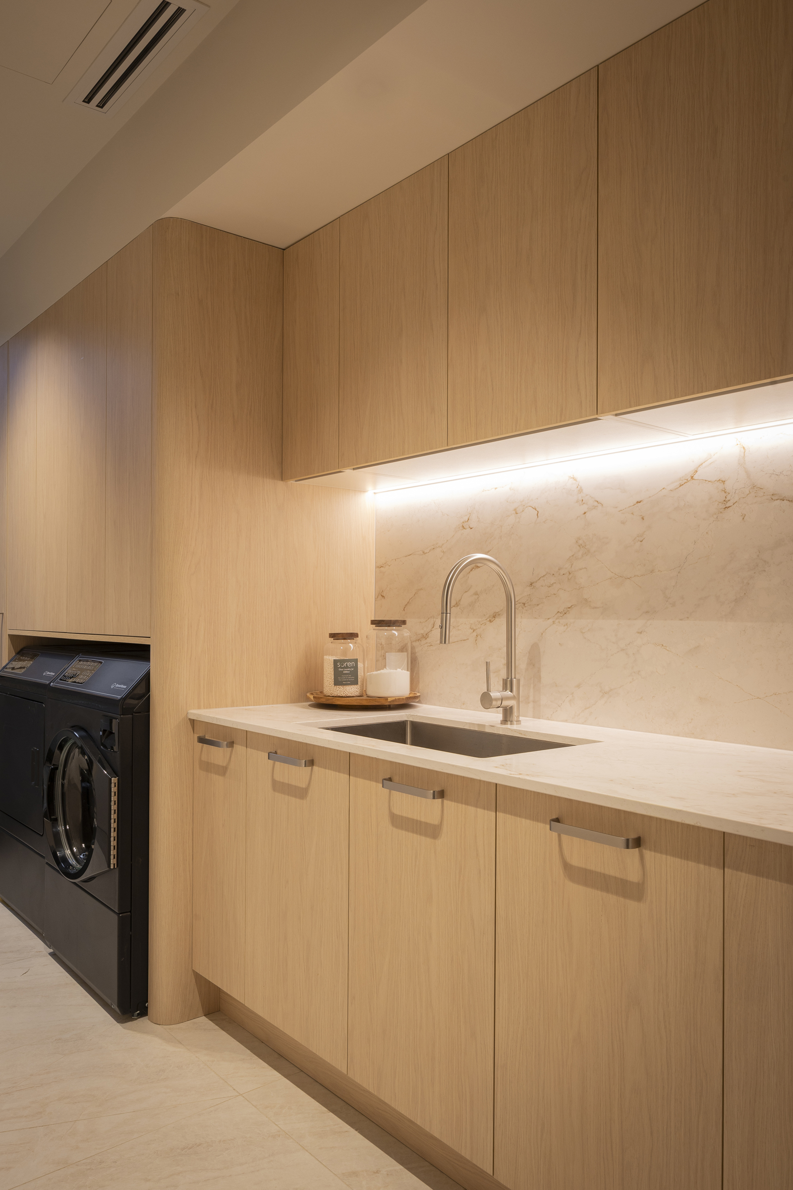 Timber look laundry with warm lighting, side by side laundry appliances, lots of storage and bench space, and a sink and tap