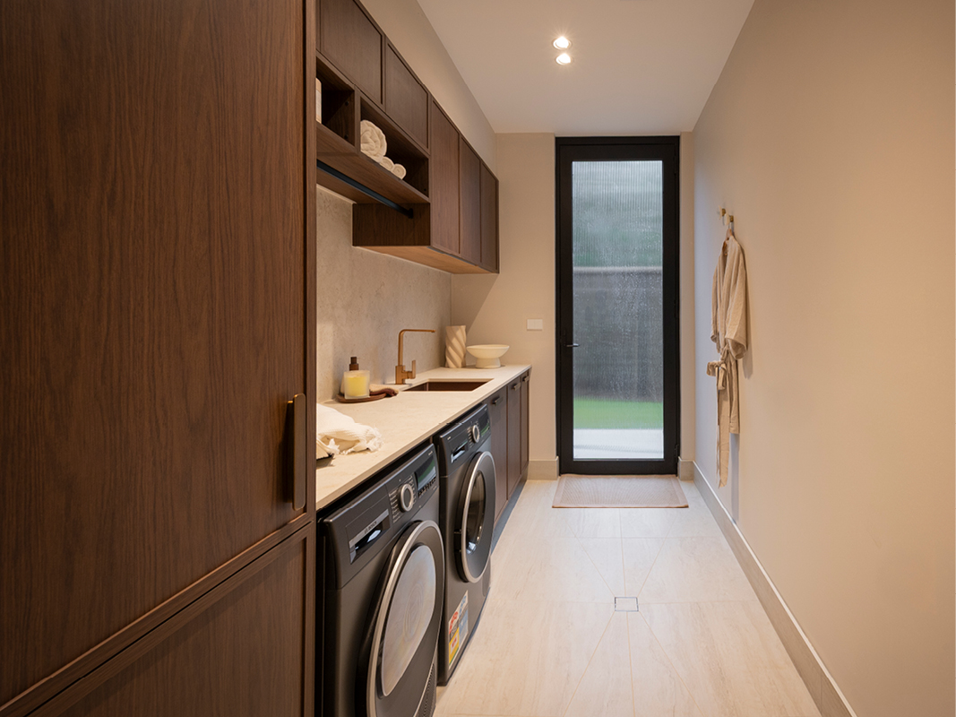 Kinsman laundry with rich walnut-toned cabinetry, open shelving, bronze tapware and dark-finish washer and dryer beside a glazed door.
