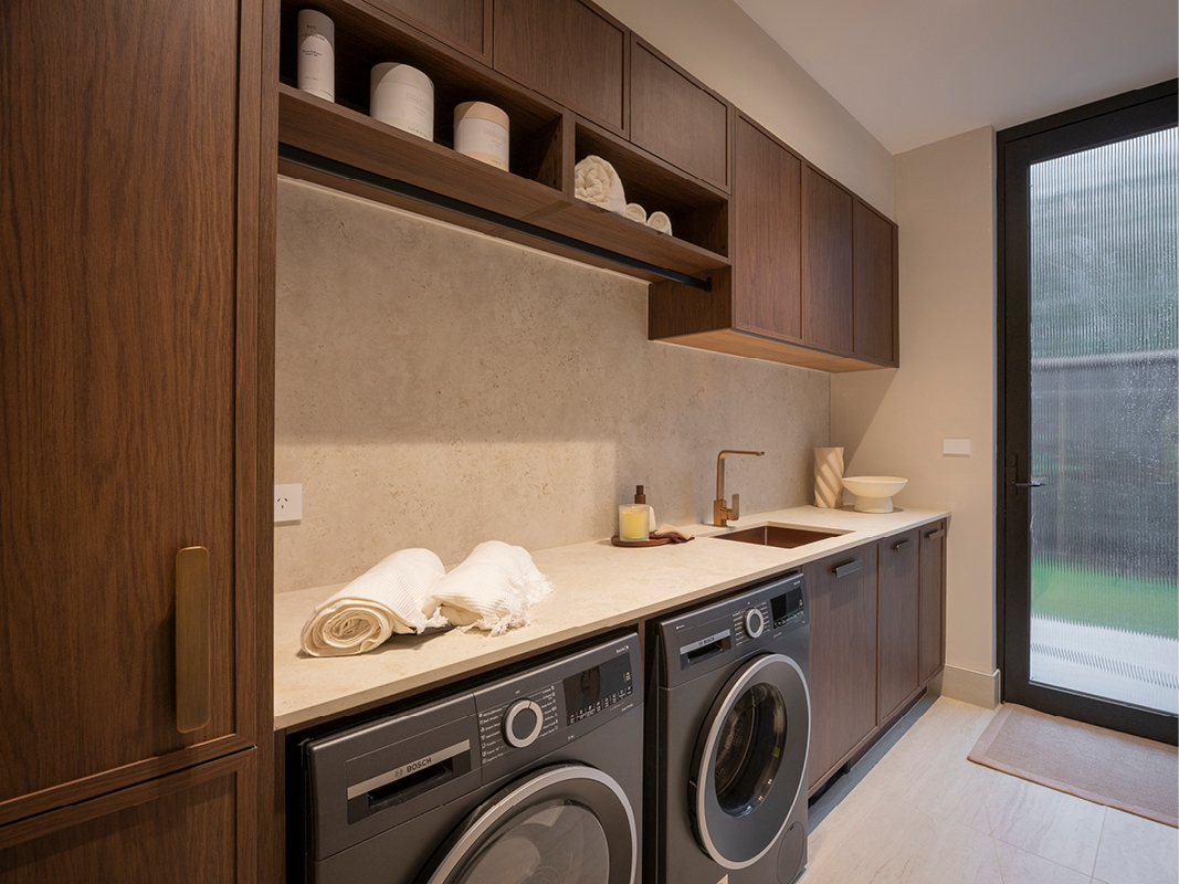 Kinsman laundry featuring open timber shelving, stone-inspired splashback, bronze tapware and side-by-side dark-finish washer and dryer.
