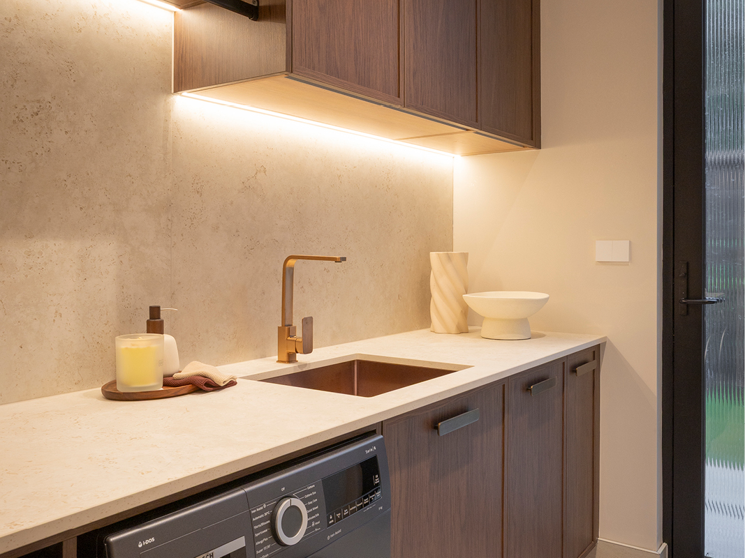 Close-up of Kinsman laundry sink in dark stone with bronze mixer tap, stone-inspired benchtop and under-cabinet lighting.