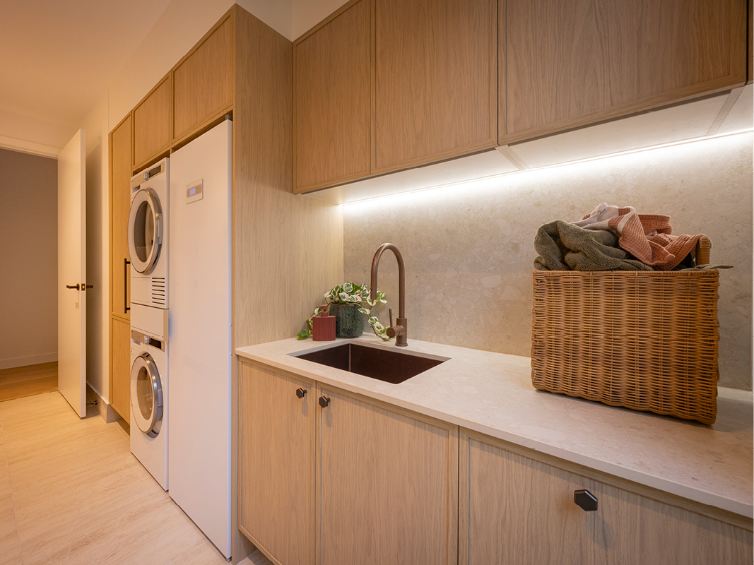 Kinsman laundry with light timber cabinetry, stacked washer and dryer, dark undermount sink, brushed copper tapware and woven storage basket.