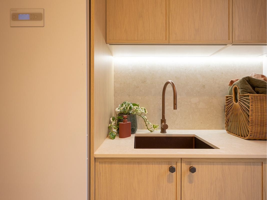 Close-up of Kinsman laundry sink in dark stone with brushed copper mixer tap, stone-inspired splashback and under-cabinet lighting.