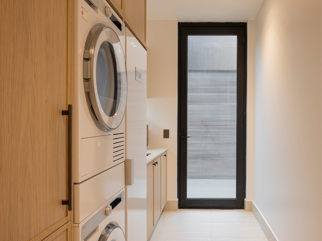 Kinsman laundry featuring stacked washer and dryer, black hardware, and light timber cabinetry beside a black-framed glazed door.