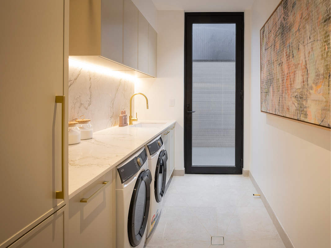 Kinsman laundry with soft greige cabinetry, brass tapware and handles, marble-look benchtop, and side-by-side washer and dryer.
