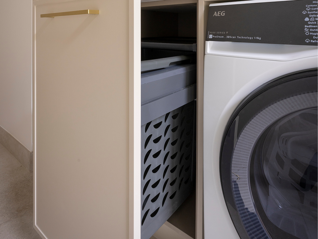 Close-up of Kinsman laundry pull-out hamper storage in soft grey, set beside cabinetry with brass handle and clothes dryer.