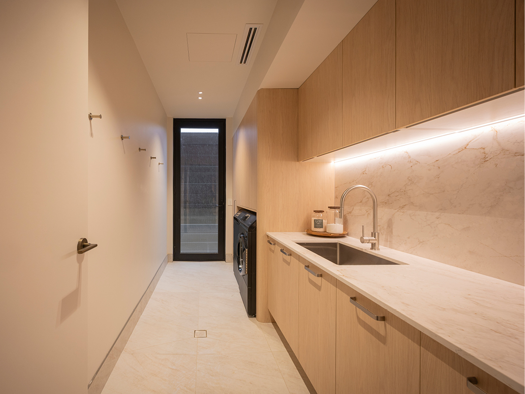 Kinsman laundry with light oak cabinetry, marble-look splashback, brushed nickel tapware and dark-finish washer beside a glazed door.