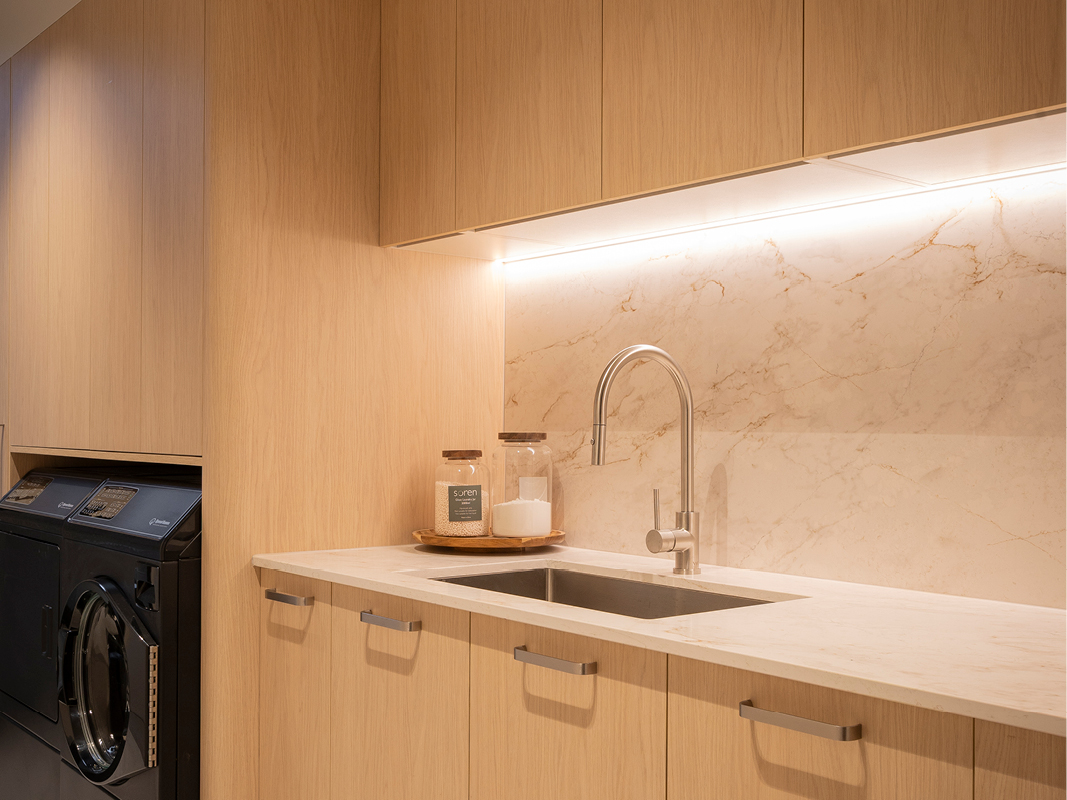 Close-up of Kinsman laundry sink in stainless steel with brushed nickel mixer tap and marble-look splashback under cabinet lighting.
