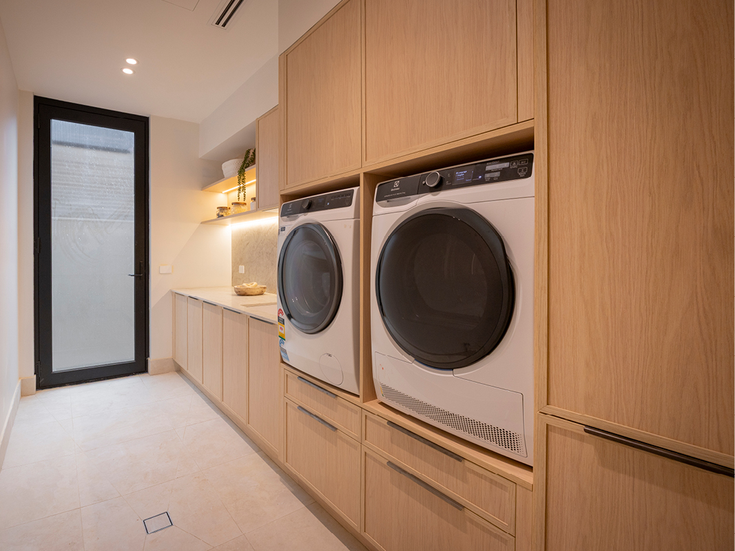 Kinsman laundry with light oak-toned cabinetry, stacked washer and dryer, and floating shelf with integrated lighting.