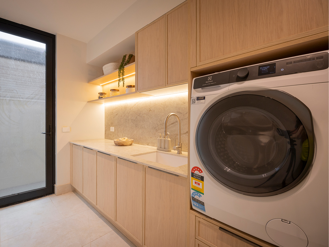 Kinsman laundry featuring light timber cabinetry, front-loading washer and dryer, and floor-to-ceiling storage beside a glazed door.